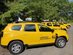 Four yellow airside operations vehicles parked on a gravel road beside some trees.