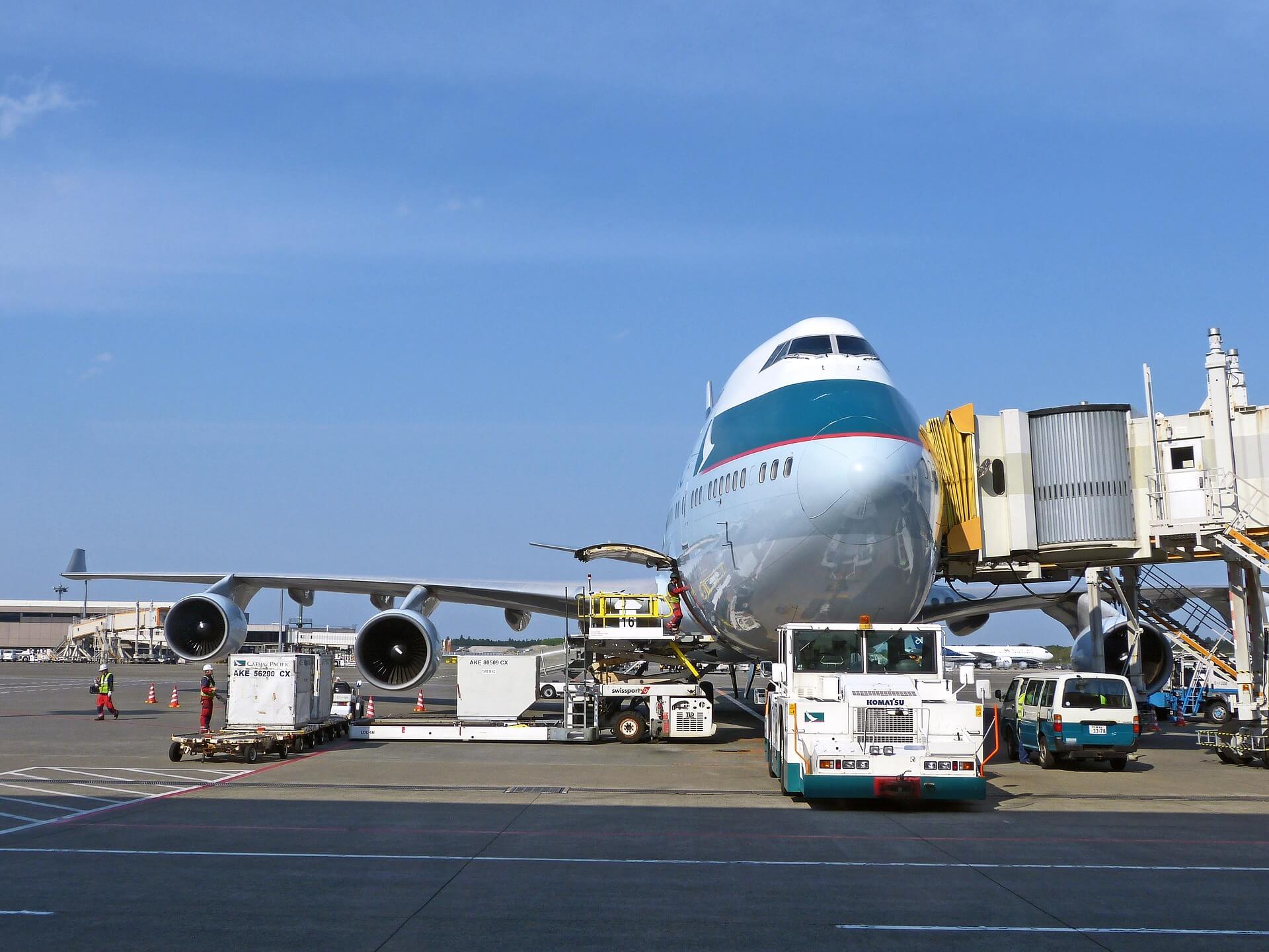 A jumbo jet plane sits on the runway apron with passenger boarding ramp and tow vehicles.