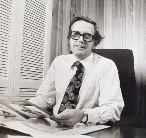 Tony Walker sits at a desk in shirt and tie reading a brochure.